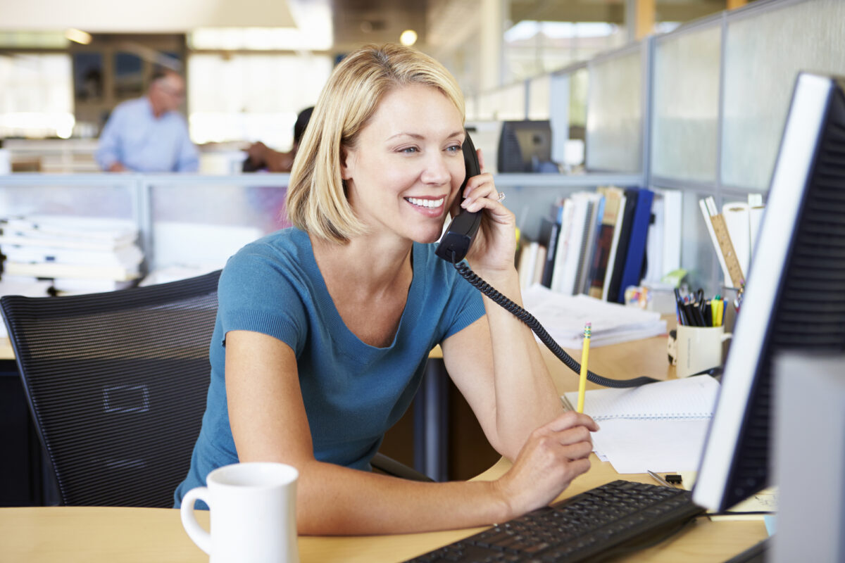 Office worker smiling while talking on a desk phone and using a computer.
