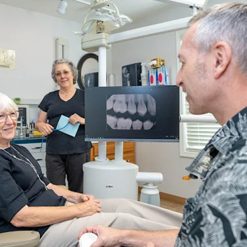 Dentist consulting with a patient while showing dental x-rays on a screen, with an assistant in the background. Dentist consulting with a patient while showing dental x-rays on a screen, with an assistant in the background.